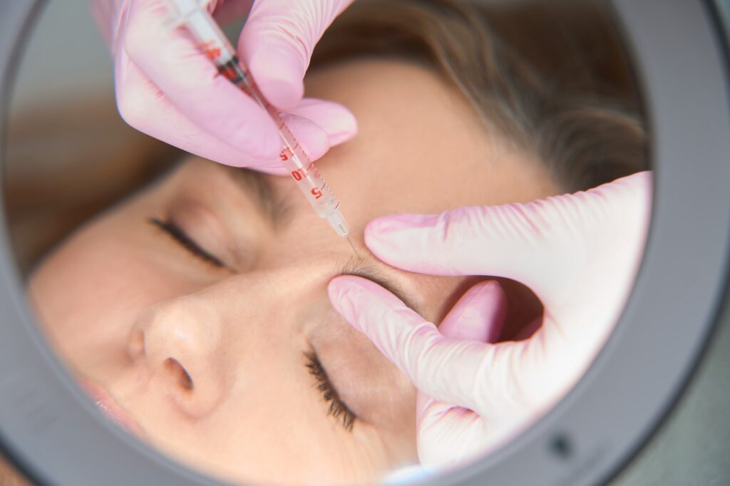 Close-up view of a professional cosmetologist wearing pink gloves administering a precise cosmetic Botox Injections to a woman's face, reflected in a magnifying mirror, depicting advanced skincare treatment.